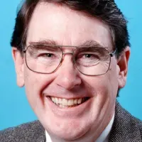 A portrait of a smiling man. He wears glasses, a suit jacket, and a tie.