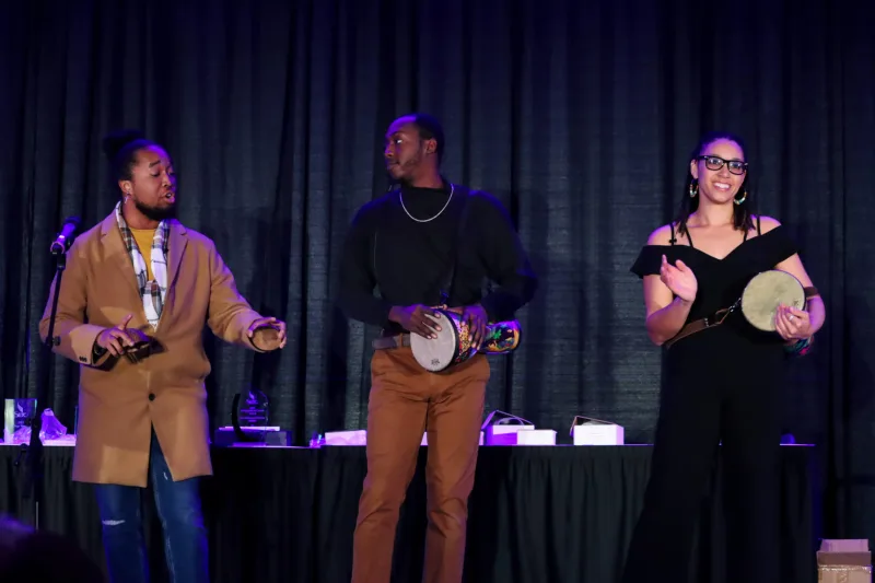 Three performers in front of a curtain. On the left, a performer is singing. In the middle, a performer is playing a hand drum. On the right, a performer is clapping.