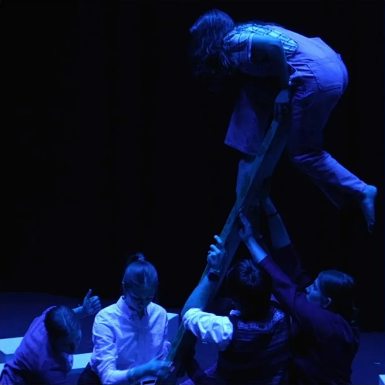 Actors bathed in blue light on a dark stage. One actor is lifted atop a ladder, with four actors are on the stage floor beneath.